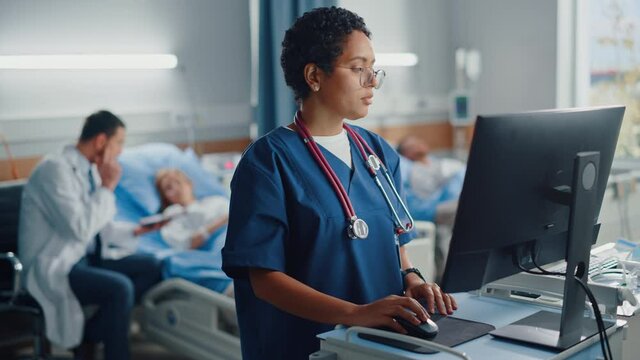 Hospital Ward: Professional Black Female Head Nurse Uses Medical Computer. In the Background Modern Equipment Clinic Doctor Consulting Patient Recovering After Successful Surgery in Bed