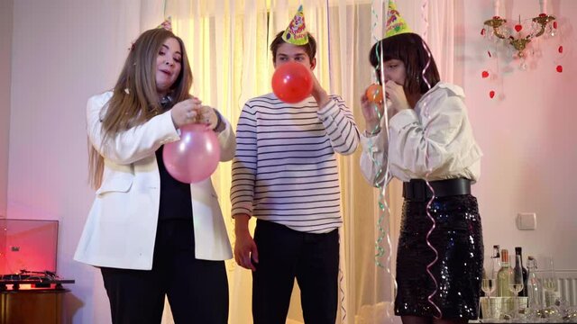 Three joyful young friends blowing balloons for birthday party talking and laughing. Positive Caucasian men and women having fun preparing partying indoors. Fun and joy concept.