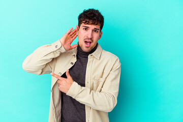 Young caucasian man isolated on blue background trying to listening a gossip.