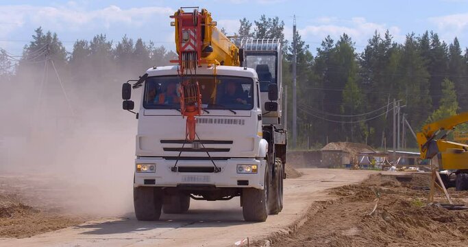 A truck crane with a yellow telescopic boom rides on a dusty road at a construction site 