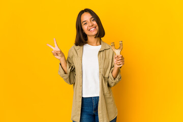 Young hispanic woman holding a slingshot joyful and carefree showing a peace symbol with fingers.