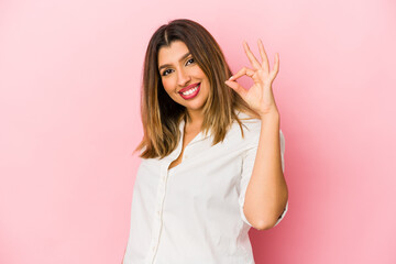 Young indian woman isolated on pink background winks an eye and holds an okay gesture with hand.