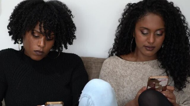 Two millennial african women sitting on sofa using mobile phones ignoring each other