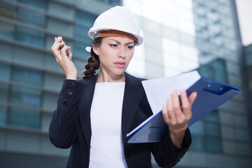 Young woman engineer in suit and hat is taking folder and discussing project by the phone near the building.