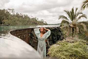 Happy young woman in long  dress stand and pose near swimming pool and enjoying summer vacation under palm trees