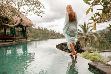 Happy young woman in long  dress walking on the edge of swimming pool and enjoying summer vacation in daytime