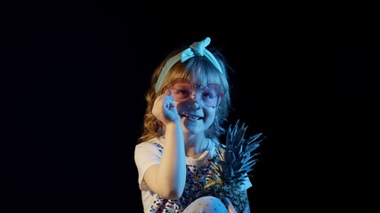 Stylish teenager caucasian kid girl with pineapple on black background smiling, fooling around. Child look at camera, making faces in night club backlit with blue neon light. Futuristic style