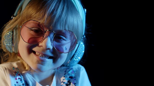 Stylish Trendy Child Kid In Sunglasses At Disco Party Cyberpunk Club Looking At Camera And Smiling. Close-up Portrait Of Young Playful Blond Girl In Bright Colorful Neon Rays Light On Black Background