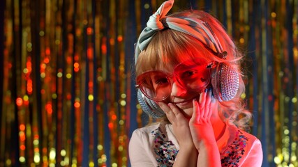 Portrait of trendy stylish girl looking at camera, smiling. Cute child kid in red neon lighting posing at disco cyberpunk club. Futuristic hipster teenager children making faces, fooling around
