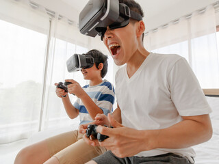 A father and son Sitting in bed. Asian family have fun and enjoy playing the game by wearing virtual glasses. On the bed in the room. Holiday concept..