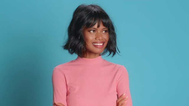 Happy pleased dark skinned Asian young woman looks on different sides with shy expression smiles broadly shows white teeth winks eye with flirt dressed in long sleeved jumper models on blue background