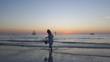 little girl with a watering can is playing at the beach during a sunset