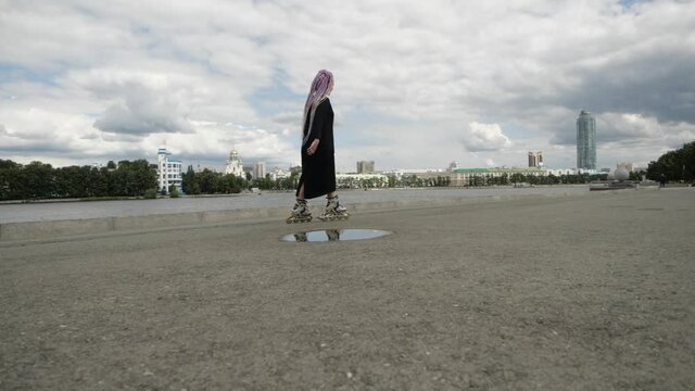 Bright young woman with colored pigtails and in a long black dress performs tricks on roller skates on the asphalt against the backdrop of the cityscape. High quality FullHD footage