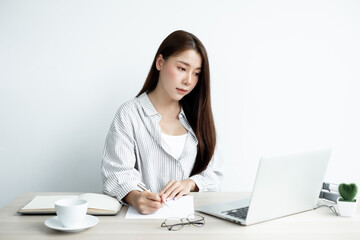 Asian female college students study online at home, enjoy extra lessons, watch videos, and take notes from a digital laptop, do homework by looking at the screen while sitting at a desk at home
