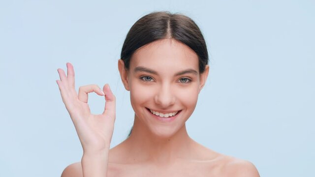 Young attractive brown-haired woman make an ok hand gesture and smile on pale blue seamless background