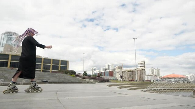 An interesting girl with colored long pigtails performs tricks on roller skates near the city fountain against the backdrop of the city landscape. High quality FullHD footage
