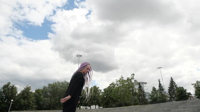 A young woman with interesting colored pigtails performs tricks on roller skates against the background of tall buildings and sky. High quality FullHD footage