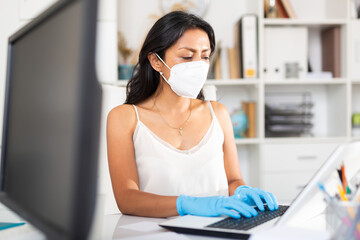 Portrait of confident Latina wearing medical mask working on laptop in office. New life reality during coronavirus pandemic