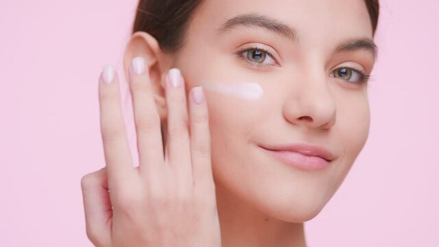Close-up portrait of young attractive beauty model with perfect skin who applies cream on her face and looks at the camera on light pink background | Skin care product advertising concept