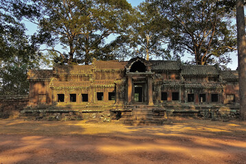 Obraz premium Ancient stone palace in Angkor. Empty window and doorways, columns at the entrance are visible. The building is dilapidated and weathered. Tropical trees against the blue sky. Cambodia