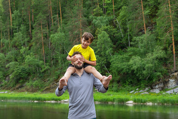 smiling man and young boy playing outdoors. boy riding on his fathers shoulders and closed his eyes with his hands