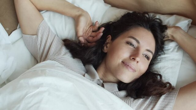 Top view POV of brown-eyed young Caucasian woman lying in bed, waking up, opening eyes, looking and smiling on camera in morning