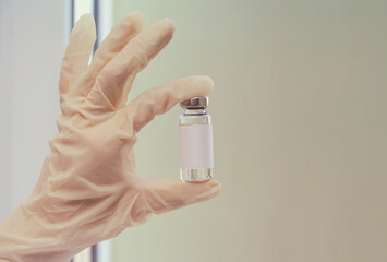 The doctor's hand in a medical rubber glove, holding a bottle of vaccine against the background of the window. Close-up, copy space, mockup for advertising. Concept of vaccination of the population.
