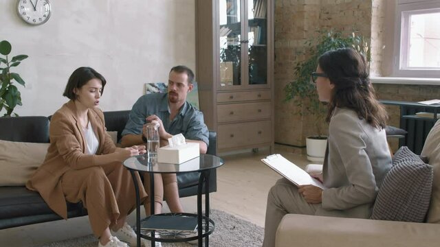 Dolly-in slowmo shot of female relational therapist with clipboard sitting in chair in her private practice office and counseling couple