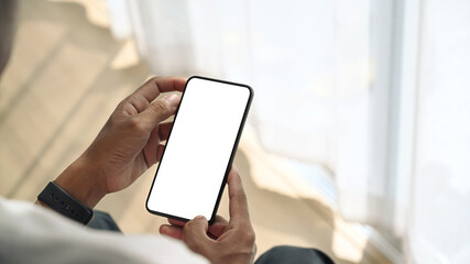 Close up view of young man sitting near window and holding mock up smart phone with white screen.