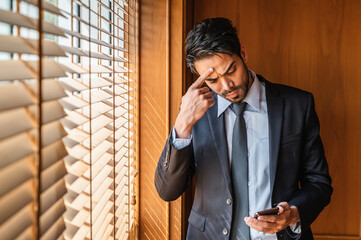 A businessman standing in front of a window with blinds in a suit feeling upset and frustrated and looking at the mobile phone in his hand.