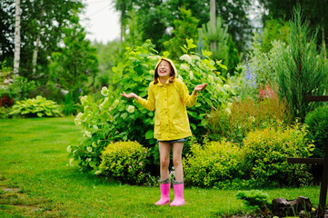 happy kid girl in yellow raincoat playing and having fun in summer garden under the rain