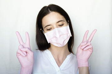 A woman in gloves and a protective mask stands on a light background in a white jacket. The concept of suppression of coronavirus