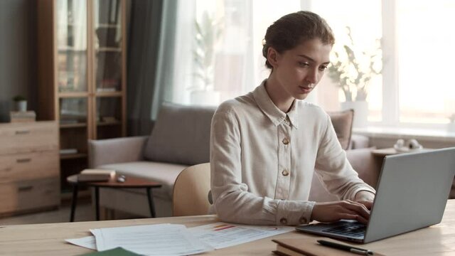Medium close-up of young professional Caucasian woman wearing formal shirt, sitting by desk in home office, typing on laptop computer, working from home
