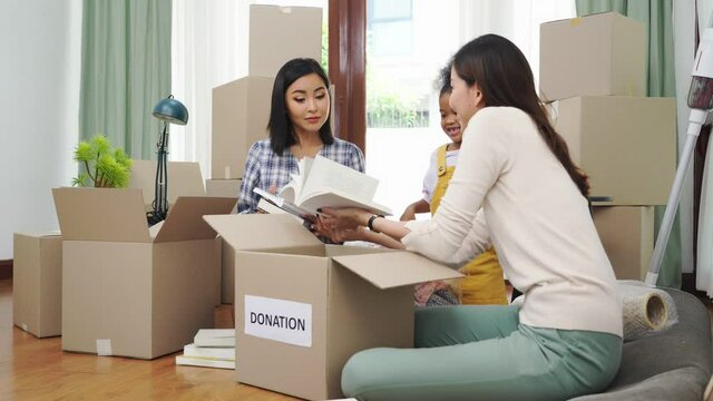 Happy young Asian lesbian couple sitting on floor surrounded by cardboard boxes selecting and organizing books into boxes while their cute African daughter standing watching and smiling next to them.