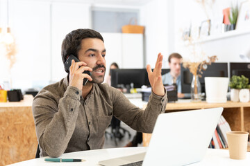 Angry hispanic man with mustache discussing on the phone in front of his laptop in the office.