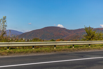 Autumn mountain landscape - yellowed and reddened autumn trees combined with green needles and blue sky on the side of a deserted road. Colorful autumn landscape scene in the Ukrainian Carpathians.