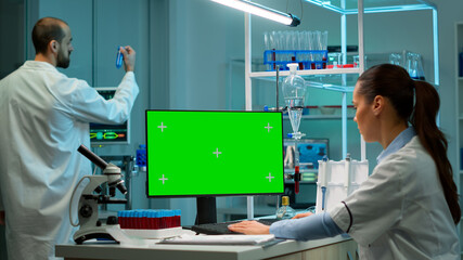 Biochemistry scientists developing drugs, female microbiologist doing research working on computer with green screen chroma key display. In background man lab researcher bringing blood samples