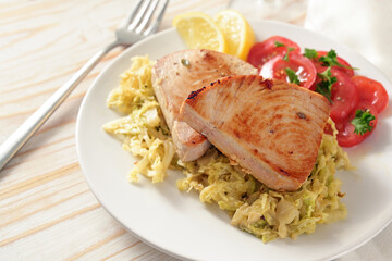 Two tuna steaks on savoy cabbage with tomatoes and lemon slices on a white plate and a light wooden table, copy space