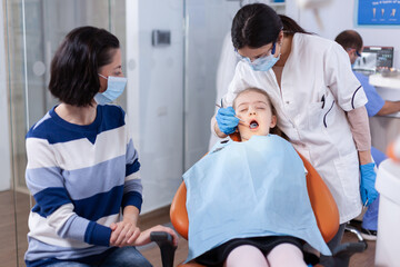 Kid wearing dental bib sitting on chair in dentistiry office with mouth open. Dentistry specialist during child cavity consultation in stomatology office using modern technology.