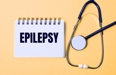 On a beige background, a stethoscope and a white notepad with the inscription EPILEPSY. Medical...