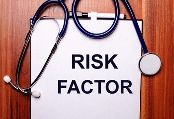 The word RISK FACTOR is written on white paper on a wooden background near a stethoscope and black-framed glasses. Medical concept