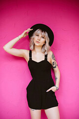 Beautiful young hipster woman with stylish blonde hairstyle, tattoos in black dress and hat posing and looking at the camera isolated on pink background.