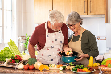 Vegetarian lifestyle. Beautiful white-haired senior couple in the kitchen prepare a vegetable soup. The chef puts chopped vegetables in the pot, on the table a mix of raw seasonal vegetables