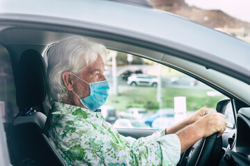 Elderly bearded man wearing surgical mask due to covid19 coronavirus ready to drive his gray car looking at the traffic in front of him - active retirees concept