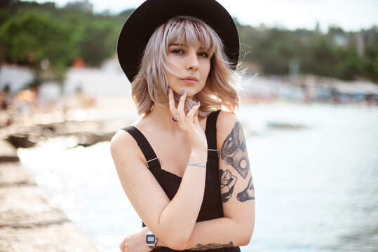 Close up portrait of blonde hipster woman in black hat and dress with tattoos and piercing outdoors