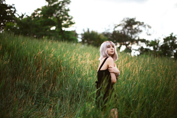 Fototapeta premium Young stylish girl with blonde hair standing in the green field in black dress, holding black hat. Portrait shot