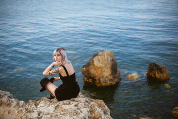 Beautiful young blonde hipster girl with tattoos wearing black dress, holding black hat sitting on...