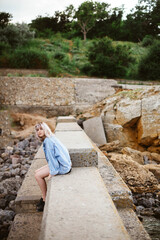 Fototapeta premium Blonde hipster girl with blonde hair in jeans jacket sitting outdoors on the rocks.