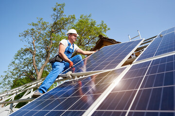 Young technician standing on metal platform installing heavy solar photo voltaic panel on blue sky...