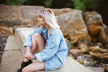 Fototapeta na wymiar Blonde hipster girl with blonde hair in jeans jacket sitting outdoors on the rocks.